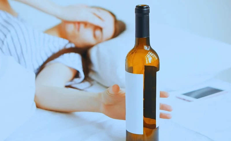 Drunk women lying on her bed reaching a bottle of wine.
